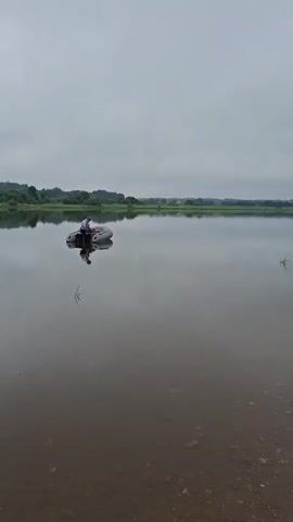 A person sits in an inflatable boat on a calm lake. The boat has a small outboard motor. The sky is overcast and the water reflects the sky and the distant treeline. The shore in the foreground is rocky.