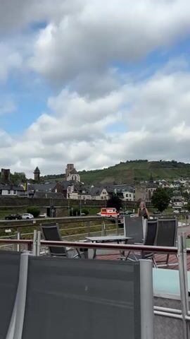The video shows a view from what appears to be a deck of a boat. In the foreground, there are grey chairs and tables, some of which are empty. Beyond the chairs, there is a railing and then a walkway or deck. In the middle ground, there is a town or village with buildings and a church spire. The town is situated on a hill that is covered with vineyards. There is a road running along the waterfront with some cars and a yellow and white vehicle that looks like an ambulance or emergency vehicle. A woman wearing a light-colored top and dark pants is walking on the walkway in the middle ground. The sky is partly cloudy with blue sky visible. The overall impression is a scenic view of a European town and countryside.