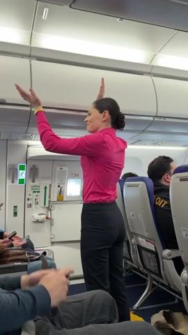 Here's a concise description of the video:

The video shows an airplane's interior, likely during boarding or before takeoff. A flight attendant, wearing a bright pink long-sleeved shirt and black pants, is standing near an emergency exit door, gesturing with her hands, possibly demonstrating safety procedures. She has her hair pulled back in a bun.  Other passengers are seated, some using their phones. The cabin has overhead compartments and standard airplane seating. One passenger has a Detroit t-shirt on.
