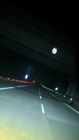 A first-person perspective video captures a nighttime drive on a highway. The road ahead is illuminated by the vehicle's headlights, revealing white lane markings and a concrete barrier on the left with red and white lights. The right side of the road also has a row of lights. A circular sign with red and blue elements is visible above the barrier. The sky is dark. The video is static, with no discernible movement except for the continuous forward motion of the road.