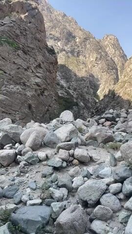 The video displays a rocky, mountainous canyon. The foreground is filled with numerous large, rounded rocks of varying sizes, interspersed with smaller stones and some sparse vegetation. The canyon walls rise steeply on either side, with sheer rock faces showing geological strata. The path ahead ascends into the distance, with more rocks and vegetation visible. The sky is clear and blue. No people or animals are present, and no actions or interactions are depicted. The scene is static and natural.