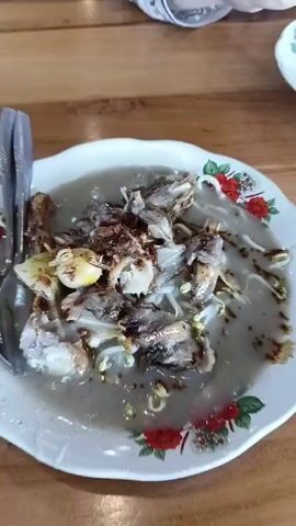 A partially eaten plate of what appears to be Soto Ayam, an Indonesian chicken soup, is shown in the video.  The soup consists of chicken pieces, bean sprouts, fried onions, and what may be boiled eggs, all in a light broth. The plate is white with a red rose pattern around the rim, sitting on a wooden table. A person's hand places a spoon and fork next to the plate. The person is wearing a white and black long-sleeved top.  The video seems to capture a meal at a restaurant or home.
