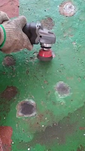 Here is a description of the video.
A person in orange work pants and a gray jacket is using a small grinder to remove rust and paint from a green metal surface. The person is wearing gloves and ear protection. There are several spots of rust on the surface.  Another grinder is visible in the background.