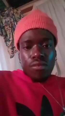 A person with dark skin is centered in the frame, wearing a pink beanie and red Adidas sweatshirt with a black logo. They have a silver chain necklace with a cross pendant, and silver hoop earrings. They look towards the left, then directly into the camera. They adjust their beanie, then briefly cover their mouth with their hand. The background includes white curtains and a corrugated metal ceiling with green lights. Upbeat music plays throughout the video.