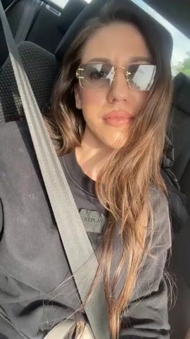 The video shows a woman in a car, wearing sunglasses and a black shirt with "REPLAY" written on it. She is wearing a seatbelt and looking at the camera. She smiles briefly at one point. There is a bag behind her on the seat. The car appears to be moving.