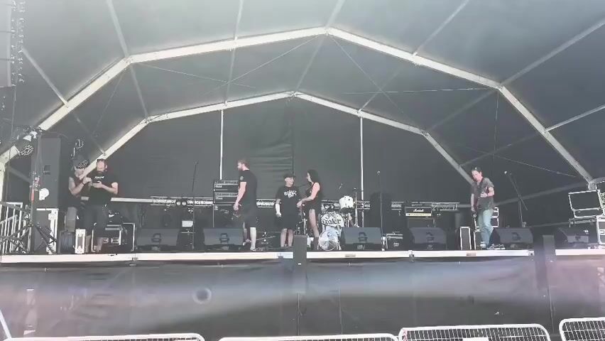 A group of five people, three men and two women, are on a stage under a large tent. Most of them are wearing black shirts. They appear to be a band preparing to perform. One man on the far left is holding a cell phone. A man in the center is holding a guitar. Another man on the far right is also holding a guitar. There is a drum set on the stage. The stage is set up with amplifiers and other musical equipment. The tent has a white, segmented roof and black fabric walls. The scene is lit by daylight, and the tent provides shade.