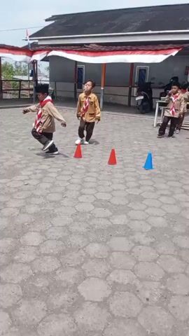 The video shows two young boys, dressed in Scout uniforms, participating in a race. The race appears to involve navigating around orange and blue cones placed on a hexagonal paved surface. The first boy, in the foreground, is running and wearing a brown uniform with a red and white scarf. He is looking forward and appears to be focused on the race. Behind him, a second boy, also in uniform, is running and appears to be holding a straw to his mouth. The background shows a building with red and white banners hanging from the roof, a motorcycle, and a table with some items on it. The overall environment suggests an outdoor event, possibly at a school or community center, on a bright, sunny day.
