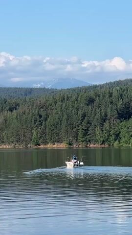 A boat with at least two people is moving across a lake. The boat appears to be a fishing boat with fishing rods visible. The water is calm and reflects the sky and trees. Behind the lake is a dense forest of green trees, and in the distance, mountains with snow-capped peaks are visible under a clear blue sky with some white clouds. The scene appears to be a sunny day in a natural, serene environment.