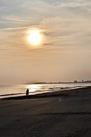The video shows a silhouetted figure on a vehicle, possibly a motorcycle or quad bike, on a beach. The sun is low in the sky, creating a bright glare over the sea and casting long reflections. The beach is mostly deserted, with a hint of vegetation in the distance. The sky is covered with thin clouds that add to the overall soft, bright lighting.