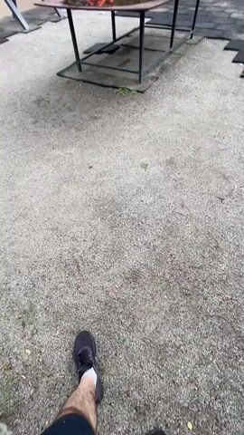 The video shows an outdoor exercise area with several pieces of workout equipment. The equipment is mostly orange and gray. In the background, there is a large apartment building with many windows. Trees and grass are also visible around the apartment building and the exercise area. The ground of the exercise area is a mix of gravel and black rubber matting. Some of the matting is unevenly laid. A rectangular metal table with a dark surface, possibly for games or other activities, is positioned in the center of the area. The sky is overcast. The camera is in motion, panning across the scene.