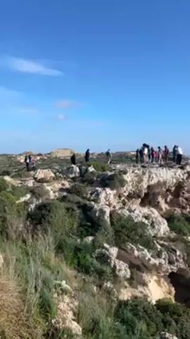 The video shows a group of people standing on a rocky cliff overlooking the ocean. The sky is blue with few clouds. The coastline is visible, with green fields and vegetation along the cliffs. The sea is a deep blue. One man stands with arms outstretched on the cliff's edge. There is a gust of wind evident.