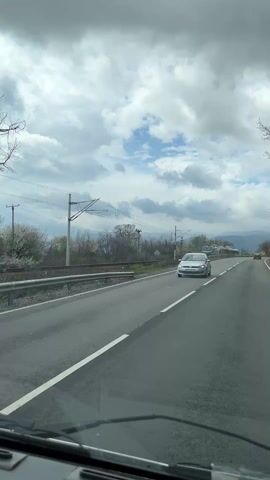 The video shows a view from a vehicle driving along a wet road next to a railway line. A green and grey train, pulling several carriages covered in graffiti, travels in the opposite direction. The sky is mostly cloudy with patches of blue. Trees and bushes line the railway. The road has a single white dividing line.