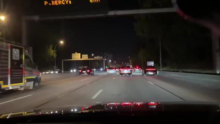 The video shows a night-time view from inside a car driving on a highway. Several other vehicles are visible on the road, including cars, a truck with emergency markings on its side, and a motorcycle. The highway has multiple lanes and is illuminated by streetlights and the headlights of passing cars. Buildings and trees can be seen in the background. The perspective suggests the video was recorded by a passenger or driver using a camera mounted on the dashboard or windshield.