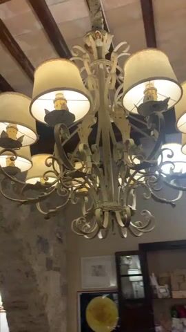 This is a video of a decorative chandelier with cream-colored lampshades hanging from a wooden beamed ceiling. The chandelier is ornate, made of metal, and appears to be antique. It has multiple arms, each holding a lamp with a shade. The lighting is warm and casts a glow. The background shows a stone wall and some artwork on the wall. There are also glimpses of windows and a spiral staircase. The video pans slightly to show different angles of the chandelier and its surroundings.