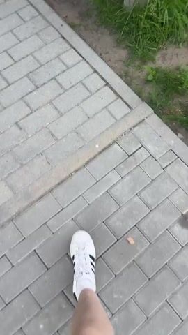 A person's legs and feet wearing white sneakers are walking on a paved path. An orange cat is lying on its back on a wooden park bench, stretching and arching its body. The bench is surrounded by green grass and small white flowers.