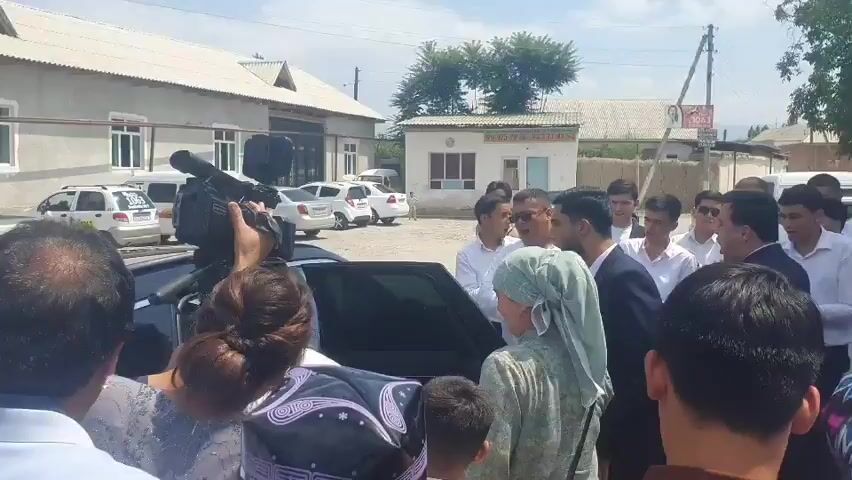 This media file captures a bustling outdoor scene with a crowd of people gathered around a car. The event appears to be a public gathering or perhaps an arrival.

**Subjects:** The primary subjects are a diverse group of people, including men, women, and children. Many are dressed formally, with men in suits and women in dresses and headscarves. Some individuals are actively engaged with the car, and one person is holding a professional video camera, suggesting media coverage. Objects include several cars parked in the background, the car at the center of the gathering, and buildings that appear to be commercial or industrial structures.

**Activity/Event:** The core activity is the interaction between the crowd and the occupants of the car. People are reaching towards the car, and there's a sense of anticipation or welcome. The presence of a videographer indicates that this event is being documented. The overall atmosphere suggests a notable occasion.

**Notable Details:** The time of day appears to be daytime, with bright sunlight indicating fair weather. The setting is an outdoor area with paved ground and buildings. The architecture of the buildings suggests a location in Central Asia, consistent with the provided location context of Rizamahalla, Uzbekistan. The emotions conveyed by the people are a mix of excitement, curiosity, and possibly respect, as seen in their engagement with the car and each other.

**Visible Text:** There is some visible text on a sign on one of the buildings in the background, which appears to be in a Cyrillic or Latin script. There is also text on the side of a white van in the background.