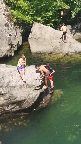 Four young men are gathered on and around a large rock adjacent to a body of water. One man, wearing purple and white patterned shorts, is seated on the rock. Two other men, one in brown shorts and the other in red shorts, are near the edge of the rock, appearing to be interacting with a third man. The third man, also in brown shorts, is bent over and appears to be either preparing to jump or being pushed into the water. In the background, a fourth man, wearing black shorts, stands on another large rock further away. The environment is lush and green with trees lining the background, suggesting a natural outdoor setting. The water is clear and green.