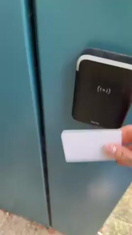 A person uses a SALTO access card to open a door. The person swipes the white card on a black access control device mounted on a blue door. The door unlocks and slides open, revealing an interior space with a striped floor mat. The person walks across the mat into the interior.