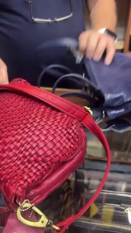 The video shows a person in a dark blue t-shirt and glasses standing behind a counter. In front of the person are two handbags, one red and one blue. The person is holding the red handbag and appears to be adjusting its strap. The red handbag is made of woven leather. The blue handbag is on the counter to the right of the red one and appears to have a textured surface. In the background, there are shelves filled with merchandise, including what appear to be wallets and other accessories. The scene suggests a retail environment, likely a shop selling handbags or leather goods.
