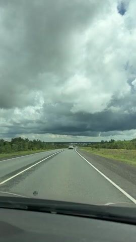 The video shows a view from inside a car driving on a wet asphalt road. The road is straight and stretches into the distance. There are two lanes in each direction, separated by white lines. Green grass and trees line both sides of the road. The sky is overcast with dark, heavy clouds, indicating potential rain. In the distance, several cars can be seen on the road. The view is from the perspective of the driver or front passenger, with the dashboard visible at the bottom of the frame.