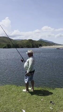 A man in a straw hat, a light-colored long-sleeved shirt, and denim shorts stands on a grassy bank near a body of water. He drops his fishing rod, then steps into the water and reaches down. The scenery includes mountains in the background, a cloudy sky, and a sandy beach visible on the opposite shore.