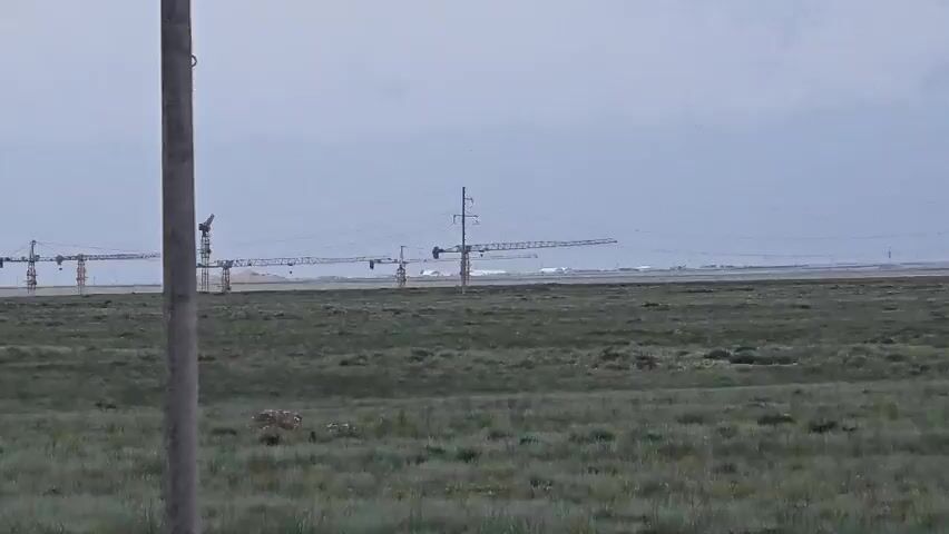 The video shows a wide, flat, grassy field under a cloudy sky. In the distance, a line of construction cranes are visible, stretching across the horizon. Power lines and transmission towers are also scattered throughout the landscape. A rough patch of earth with some debris is visible in the foreground, and a vertical pole, possibly part of a fence or utility structure, is on the right side of the frame. The scene appears to be a remote or undeveloped area undergoing construction.