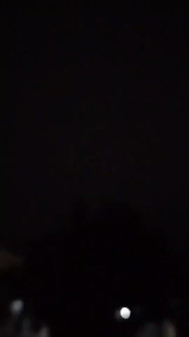 The video is extremely dark and blurry, making it difficult to discern any clear details. It appears to be filmed at night. In the bottom left corner, there is a hint of a structure, possibly a building or a wall, with a faint light source within it. The rest of the frame is predominantly black, with some very indistinct shapes that could be foliage or other objects in the darkness. There is no discernible action or interaction taking place. The overall impression is of a very low-light environment, possibly outdoors.