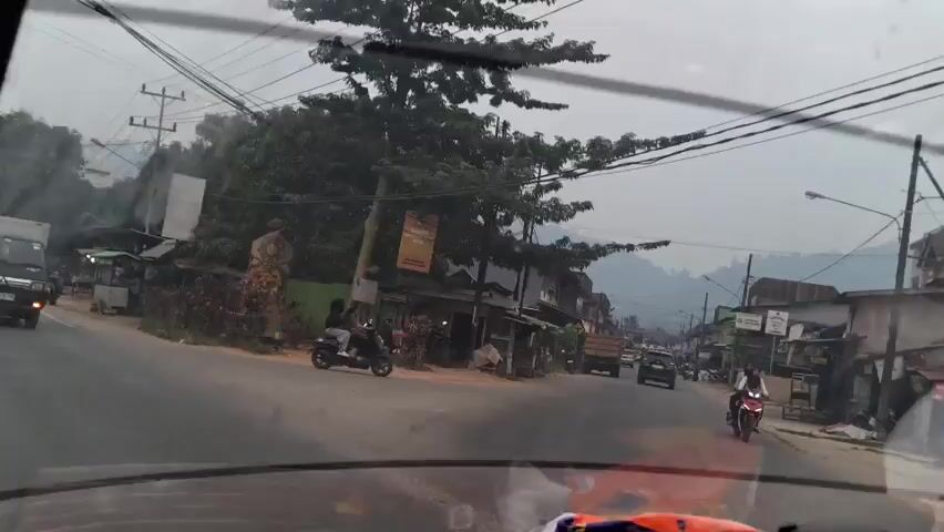 The video shows a view from inside a vehicle moving down a street.  On the left side of the street, there are several small shops and a scooter parked.  A truck is also visible further down the road.  On the right side of the street, there are more buildings and shops.  Several motorcycles are on the road, with one rider in the foreground on the right.  The sky is overcast, and there are hills in the background.  Power lines are visible strung across the street.