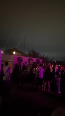 The video shows a nighttime outdoor gathering of people, likely at a party or concert.  Bright pink lights are visible, illuminating a stage or performance area where several individuals are present. A crowd of people is gathered in front of the stage, with many appearing to be dancing or socializing. The sky is dark, suggesting it is evening or night, and some clouds are visible. The overall atmosphere appears energetic and lively.