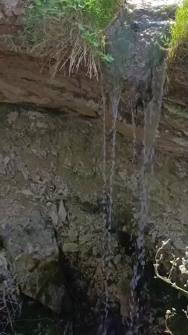 A close-up video captures a small waterfall cascading down a rocky cliff face. Green grass and foliage sprout from the top of the cliff, providing the source of the water. The water streams down the textured, grey and brown rock, eventually disappearing into the shadows at the bottom of the frame. A few dry plant stems are visible in the lower right corner of the image.
