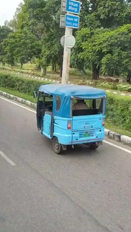 A light blue three-wheeled auto rickshaw with a blue canopy drives away on a paved road. A person is visible in the driver's seat. The auto rickshaw has a green license plate. The road is bordered by a white line on the left and grass and trees on the right. A utility pole is visible on the left side of the road. In the background, there are more trees and a tractor.