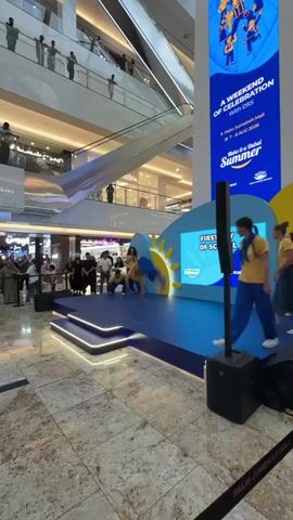 A video shows a group of young performers on a stage in a mall. The performers, dressed in yellow shirts and blue pants, are dancing. The stage is decorated with blue and yellow accents, and a backdrop with the words "First Day of School" is visible. A large banner with an image of children and text advertising "A Weekend of Celebration" is also displayed. Several people are watching the performance from the audience. The mall environment includes escalators and shops in the background.