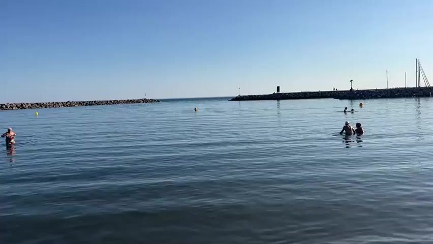 The video opens with a wide shot of a calm sea under a clear blue sky. In the distance, a breakwater extends into the water, with a lighthouse or beacon at its end. A few people are visible swimming and wading in the water. To the right, a breakwater also extends into the sea, and there are sailboats docked in a harbor. The camera pans to the right, revealing a sandy beach with several people sunbathing and playing. A child is seen walking into the water. Further down the beach, more people are gathered, some under a beach umbrella. Buildings are visible in the background, suggesting a coastal town or resort. The sun is shining brightly, casting a warm glow on the scene. The video ends with a shot of the beach and the distant buildings.