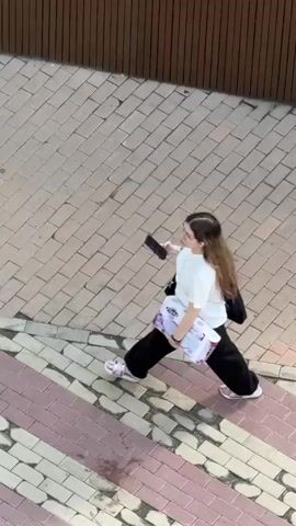 A young woman walks across a brick-paved area, holding a smartphone in her right hand and a package of tissues in her left arm. She is wearing a white t-shirt, black pants, and white sandals. A black bag is slung over her shoulder. The camera angle is a high-angle shot looking down, tracking her movement across the patterned brick. The surrounding environment is a brick pathway with adjacent architectural features partially visible.