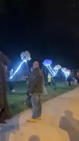 At night, a sidewalk leads to a grassy area, where several light displays resembling swings are set up. Each display features a metal frame and a seat adorned with LED lights that form an arc leading up to a fluffy, illuminated cloud. The clouds are lit in various colors, including white, blue, and green. People are scattered around the displays: a man in a green jacket and yellow shoes stands on the sidewalk, observing the scene. A woman in a similar green jacket and gray pants walks by, holding her phone, possibly taking pictures. Children and adults are sitting on the illuminated swings, enjoying the ride. The environment appears to be a park or outdoor exhibition space. The overall atmosphere is festive and cheerful.
