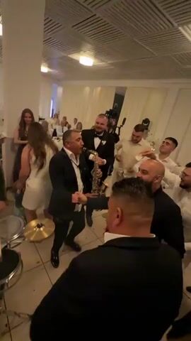 The video captures a lively scene at what appears to be a wedding reception or similar celebration. A band is performing, with a man playing the saxophone and another on drums.  Several other men surround them, clapping and enjoying the music. There are also numerous guests in the background, mingling and dancing. The room is decorated with white curtains, pillars, and what looks like modern lighting. The atmosphere is festive.  A cameraman is also visible, filming the event. The lighting changes from white to blue at one point in the video.