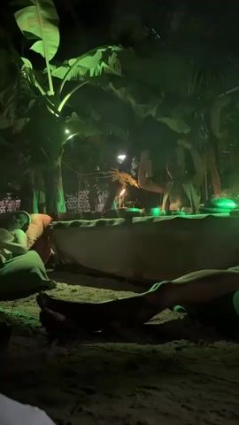 The video captures a nighttime scene in Mũi Né, Vietnam, with a group of people gathered outdoors. The setting appears to be a beach or a sandy area, surrounded by lush tropical foliage, including large banana leaves and palm trees, illuminated by vibrant green lights.

In the center of the frame, a person stands behind a large, table-like structure covered with a light-colored cloth. This individual is actively engaged, holding up what appears to be a small object, possibly for decoration or ceremony, and gesturing with their arms. A small flame flickers on the table, adding to the ambiance. Around the table, other individuals are seated or lounging on the sand, some appearing relaxed and engaged with the proceedings. Their legs are visible in the foreground, resting on the sand.

The overall mood is serene and possibly ceremonial or celebratory, enhanced by the dim lighting and the natural surroundings. The green lighting creates an ethereal and intimate atmosphere. The time of day is clearly night, indicated by the darkness and the artificial lighting. There is no visible text in the media.