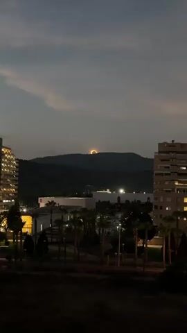 A video taken from a high vantage point shows a cityscape at dusk. The sky is a dusky blue with some wispy clouds. In the distance, a mountain range is silhouetted against the sky. On top of the mountain, a bright orange, circular object is visible, partially obscured by the mountain's peak. The object appears to be the sun setting or rising behind the mountain, casting a warm glow.

In the foreground, several buildings are visible. On the left, a tall, modern building with many windows is illuminated from within. On the right, another large building with multiple balconies and windows is also lit. Between these buildings, a lower building with a white facade and several windows is visible, along with a cluster of palm trees. The area in front of the buildings appears to be a park or courtyard, with streetlights casting pools of light on the ground. The overall impression is one of a peaceful evening in an urban or suburban setting, with the striking natural phenomenon of the sun's setting or rising behind the mountains providing a dramatic focal point. There are no people visible in the video.