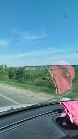 The video is filmed from inside a car looking out the windshield. The sky is blue with some scattered clouds. The car is on a paved road with a white line on the right side. The road goes through a rural area with green trees and fields. There are some hills in the distance. A pink piece of fabric, possibly underwear, is hanging on the inside of the windshield on the right side. The fabric is translucent and appears to be soiled. The dashboard of the car is visible at the bottom of the frame. There are two other cars visible on the road ahead. One is black and appears to be a sedan, the other is smaller and further away.