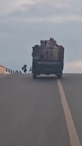 A black Mitsubishi pickup truck is driving on a paved road. The truck bed is loaded with at least eight people, who appear to be women, sitting down. They are all wearing headscarves or hijabs in various colors, including white, beige, light blue, and red. The truck has "MITSUBISHI" written on the tailgate. The road has a yellow line down the center. On the left side of the road, there is a bridge with a railing. In the background, there are trees and palm trees, and the sky is overcast. The video appears to have been filmed from another vehicle following behind the truck.
