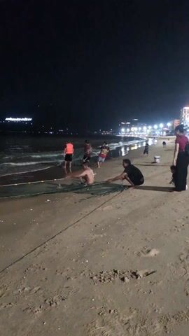 At night, on a sandy beach illuminated by distant city lights, a group of people are engaged in fishing with a large net. One man is lying in the water, partially submerged, handling the net. Another man crouches on the shore, also tending to the net. Other individuals stand nearby, some wading in the shallow water, seemingly assisting with the fishing activity. A woman with a child and a backpack stands further back on the beach, observing the scene. The environment is dark with the ocean waves lapping gently against the shore. A few buckets are visible on the sand near the group.