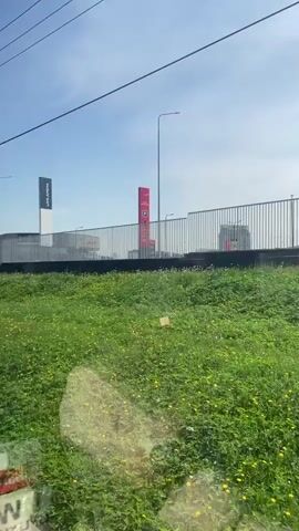 The video shows a view from a moving train. A field of green grass and small yellow flowers fills the foreground. Beyond the field is a white metal fence bordering a road above. Cars are visible moving along the road, and above the fence are signs for "AVIVA ARENA" and "ШУУМЕН". Electrical lines run overhead against a bright blue sky.