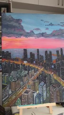 This media file is a close-up, eye-level shot of an acrylic painting on canvas, displayed on a wooden easel. The painting depicts a cityscape at sunset, viewed from an elevated perspective. The sky is a dramatic gradient of deep blue at the top, transitioning through vibrant pinks and oranges near the horizon, with dark, textured clouds in the upper left. Below the sky, a dense collection of city buildings is rendered, predominantly in dark tones, with some illuminated windows suggesting nighttime. Streaks of yellow and orange light indicate car headlights and taillights on roadways weaving through the urban landscape. The foreground features a cluster of smaller buildings with more varied colors and textures. The overall mood of the painting is atmospheric and captures the transition from day to night in a bustling city. There is no visible text in the image.