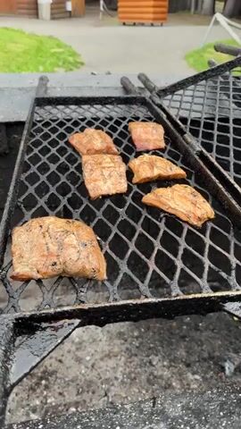 The video shows a close-up shot of several pieces of raw meat on a black metal grill. The grill is positioned over a bed of hot embers and burning logs, indicating an outdoor barbecue. In the background, out of focus, are glimpses of grassy areas and wooden structures, possibly a lodge or cabins. The meat appears to be seasoned and is beginning to cook, with some pieces showing grill marks. The overall impression is of food preparation in a rustic, outdoor setting.