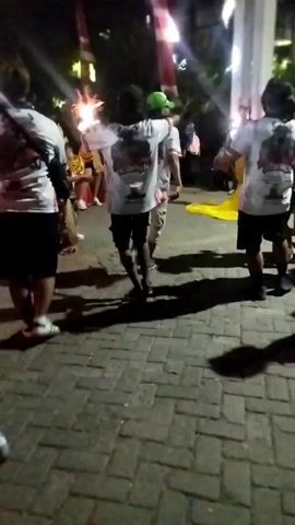 A group of people are visible at night. Most are wearing white t-shirts and dark shorts. Some are dancing. There are flags and lights in the background. The ground is paved with brick-like stones. The image is blurry and low quality.