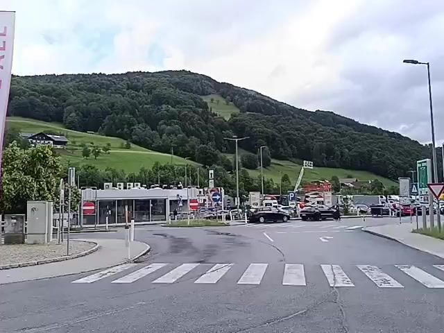A video shows a street scene in front of a building with a mountain in the background. There are three large banners on the left side of the video. The banners have text on them. The first banner has "20" and "BIRTHDAY!" on it. The second banner has "SONG CONTEST VIENNA 2026" on it. The third banner has "WE LOVE ALL" on it. There is a parking sign with a blue "P" on it and a symbol of a car with a line through it. There are several cars and a bus in the video. A man in a light-colored shirt and dark pants is walking towards the left side of the video. There are trees and grass on the mountain in the background. The sky is cloudy.