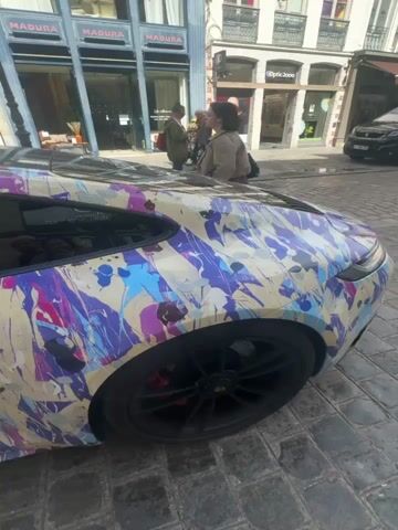 The video showcases a distinctive car, resembling a Porsche, parked on a cobblestone street in a bustling urban environment. The car is immediately eye-catching due to its unique, multi-colored splatter paint job. The base color appears to be a light cream or beige, overlaid with abstract splashes and drips of purple, various shades of blue, pink/magenta, white, and black. The car features black wheels with visible red brake calipers.

The setting is a European city street, characterized by traditional architecture and cobblestone paving. Several shopfronts line the street in the background. Visible signage includes "DESSANGE PARIS" (a hair salon), "MADURA" (a home decor store), and "Optic 2000" (an optician). These details and the overall ambiance are consistent with the provided location context of Lille, France.

Throughout the video, pedestrians are seen walking past the parked car and along the street. The weather appears sunny, casting clear shadows, indicating it is daytime. The camera moves around the car, capturing views from its rear side to its front, highlighting the artistic paintwork and the surrounding street scene. The car itself is stationary, with no specific activity taking place other than being on display.