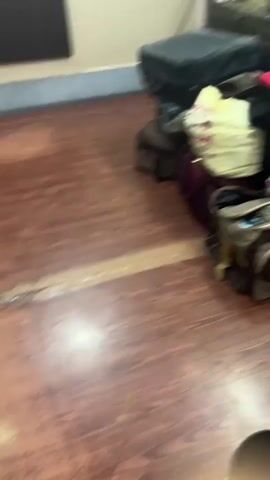 The video shows a room with a pile of luggage in the foreground. The luggage is made up of suitcases, bags, and other travel items. In the background, there is a door, a window, and a wall with a light fixture. The room appears to be furnished, but the furniture is not clearly visible. The lighting in the room is dim, and the video is somewhat blurry. The video is short, lasting only a few seconds.