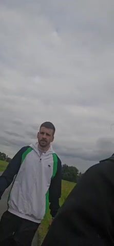 Two individuals, a man and a child, walk along a paved path. The man is wearing a white, blue, and green jacket with black pants and white sneakers. The child, a young girl, is wearing a purple jacket and holding the man's hand. A third person, a woman with red hair, is partially visible in the foreground, facing the camera. The environment appears to be a grassy field under an overcast sky. The woman speaks to the camera.