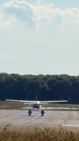 A small propeller plane is on a runway, facing away from the camera. The plane's wheels are down and it appears to be accelerating. Two faint objects are visible in the sky above and behind the plane. The runway is cracked and surrounded by dry grass and a dense line of trees in the background. The sky is clear with scattered clouds.